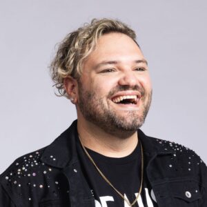 A cheerful man with curly blonde hair, wearing a black studded jacket and a gold necklace, laughing against a gray background.