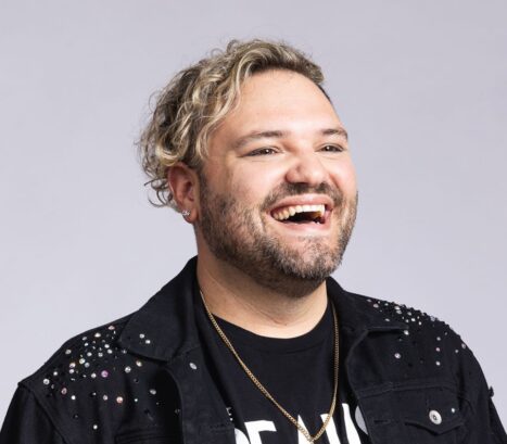 A cheerful man with curly blonde hair, wearing a black studded jacket and a gold necklace, laughing against a gray background.