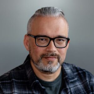 Adult man with glasses and a plaid shirt, posing for a portrait.