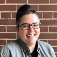 A woman with glasses smiling in front of a brick wall.