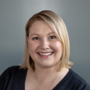 A woman with short blonde hair smiles at the camera, wearing a black top against a gray background.