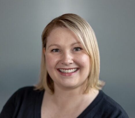 A woman with short blonde hair smiles at the camera, wearing a black top against a gray background.