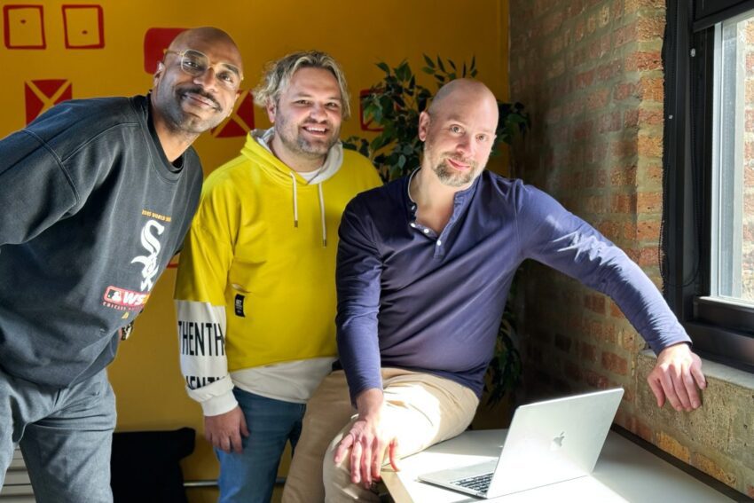 Three Orbiteers, Mo, Kyle, and Brian, posing together by a window in a room with a brick wall, with a laptop on the window sill.