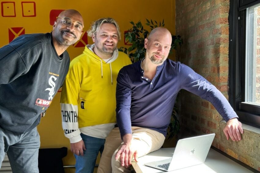 Three Orbiteers, Mo, Kyle, and Brian, posing together by a window in a room with a brick wall, with a laptop on the window sill.