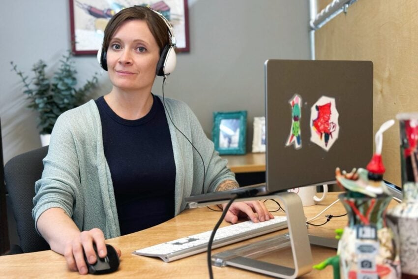Person wearing headphones, sitting at a desk with a computer, using a mouse, and looking towards the camera. Office setting with framed pictures and plants in the background.