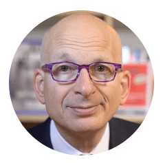 A bald man with purple glasses, wearing a suit, is seated indoors with blurred bookshelves in the background.