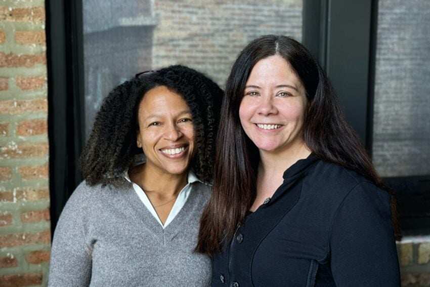 Two women stand side by side smiling in front of a window with a brick wall background.