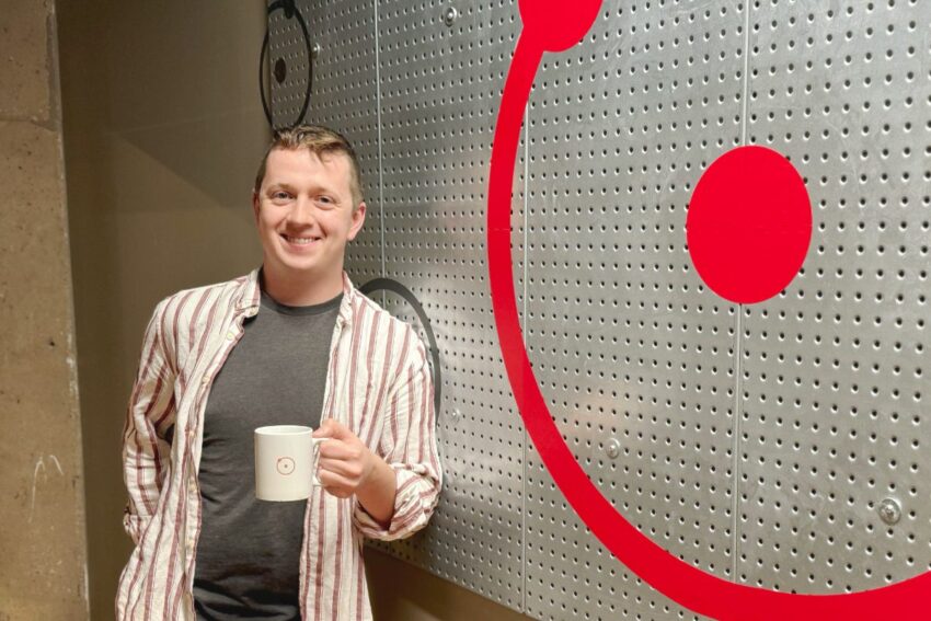 A person stands smiling while holding a coffee mug, in front of a perforated metal wall with a red circular design. They wear a striped shirt over a dark t-shirt.