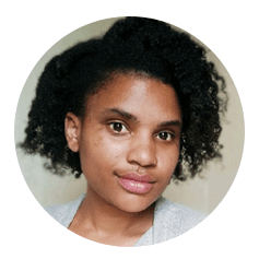 A person with short, curly hair and a neutral expression. They are wearing a gray top and are set against a light background.