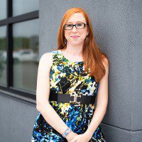 A woman with long red hair, wearing glasses and a colorful dress, stands next to a gray wall in an outdoor setting.
