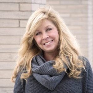 A woman with long blonde hair, wearing a dark grey sweater with a thick scarf, stands against a light brick wall, smiling at the camera.
