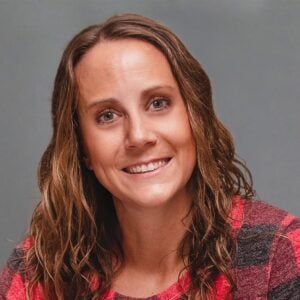 A woman with shoulder-length brown hair, smiling and wearing a red plaid shirt, is set against a plain gray background.