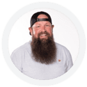A man with a long beard and a black cap smiles while wearing a light grey T-shirt against a white background.