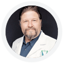 A man with long hair and a beard, dressed in a white lab coat, smiles at the camera.