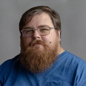 A bearded man with glasses wearing a blue shirt is looking at the camera against a plain background.