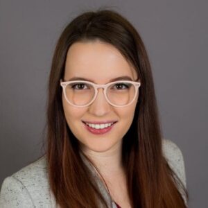 A woman with long, straight hair and glasses smiles while wearing a light gray blazer against a plain gray background.