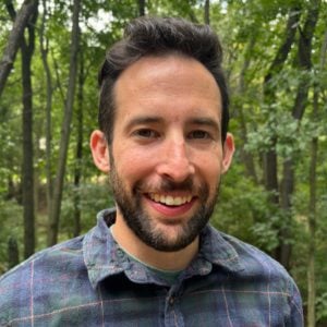 A man with a beard and a plaid shirt smiles, standing in a wooded outdoor setting.