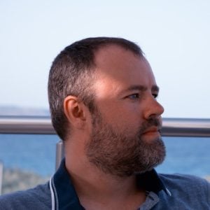 A man with a beard, short hair, and wearing a collared shirt looks to the right. A railing and ocean are visible in the background.