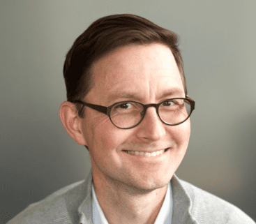 Man with glasses and a light sweater smiles in front of a gray background.