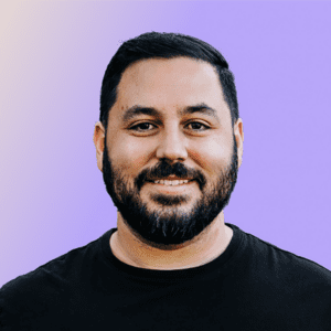 A man with a beard and short hair, wearing a black t-shirt, smiles in front of a gradient background.