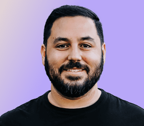 A man with a beard and short hair, wearing a black t-shirt, smiles in front of a gradient background.