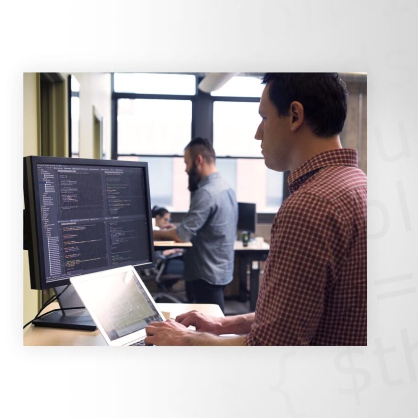 Man in a checkered shirt working on a laptop connected to a large monitor displaying code, with a person in the background working at a desk.