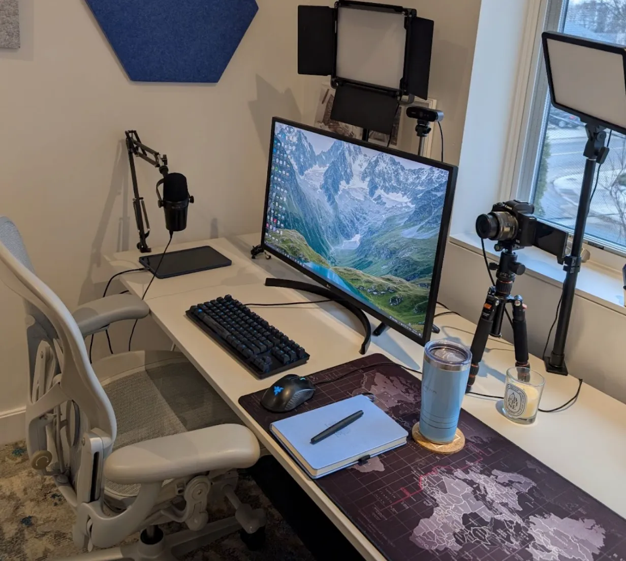 A tidy computer desk setup with a curved monitor, keyboard, mouse, microphone, camera on a tripod, lighting panels, and a drink tumbler near a window.