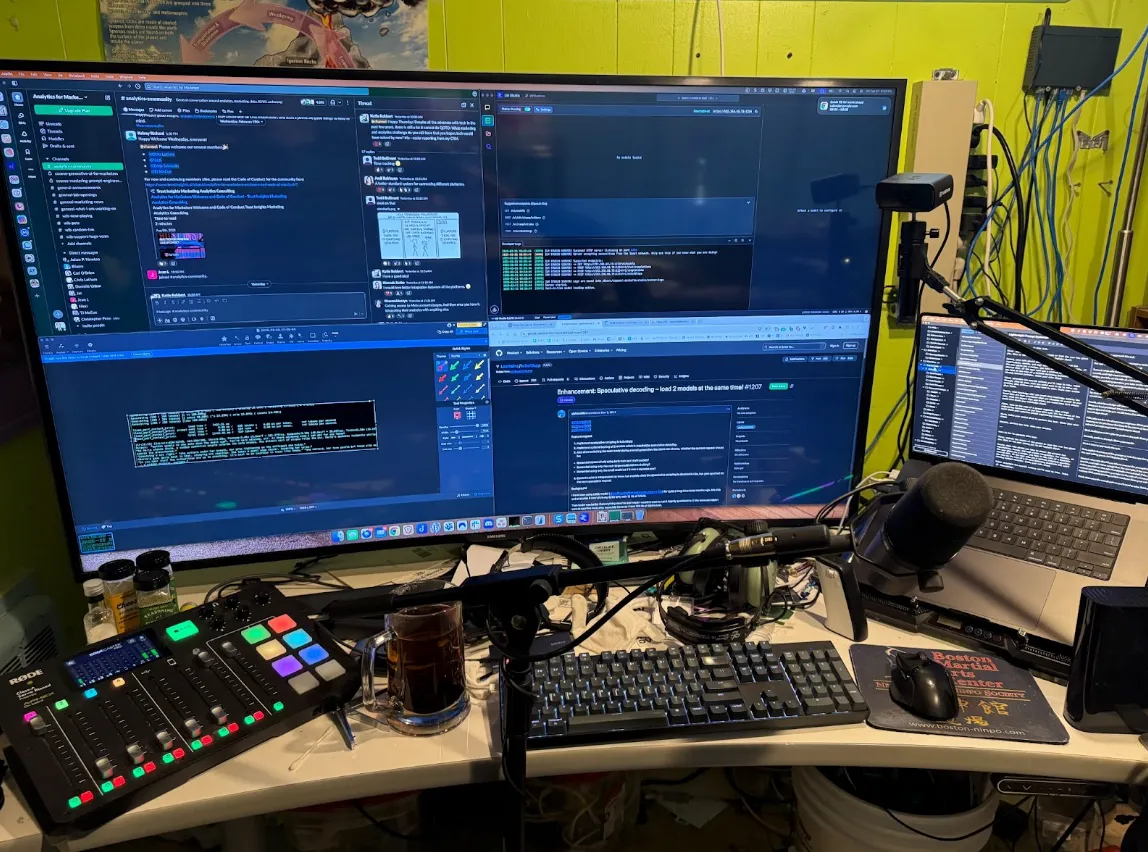 A cluttered computer workstation with multiple monitors displaying code and chat apps, microphones, audio equipment, a keyboard, mouse, and a mug on the desk.