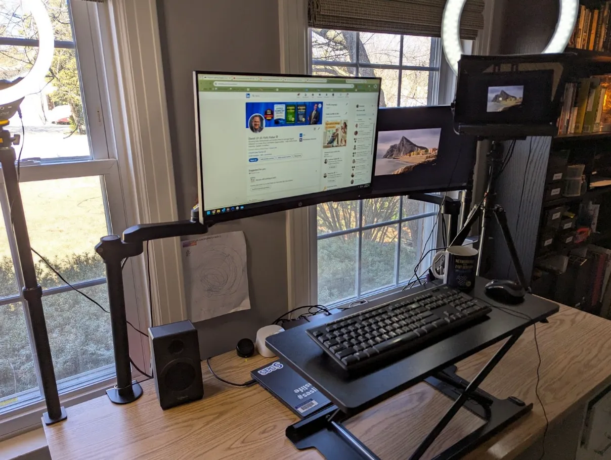 Home office setup with a computer monitor, keyboard, adjustable desk, ring lights, a small tripod with a tablet, speakers, and windows in the background.