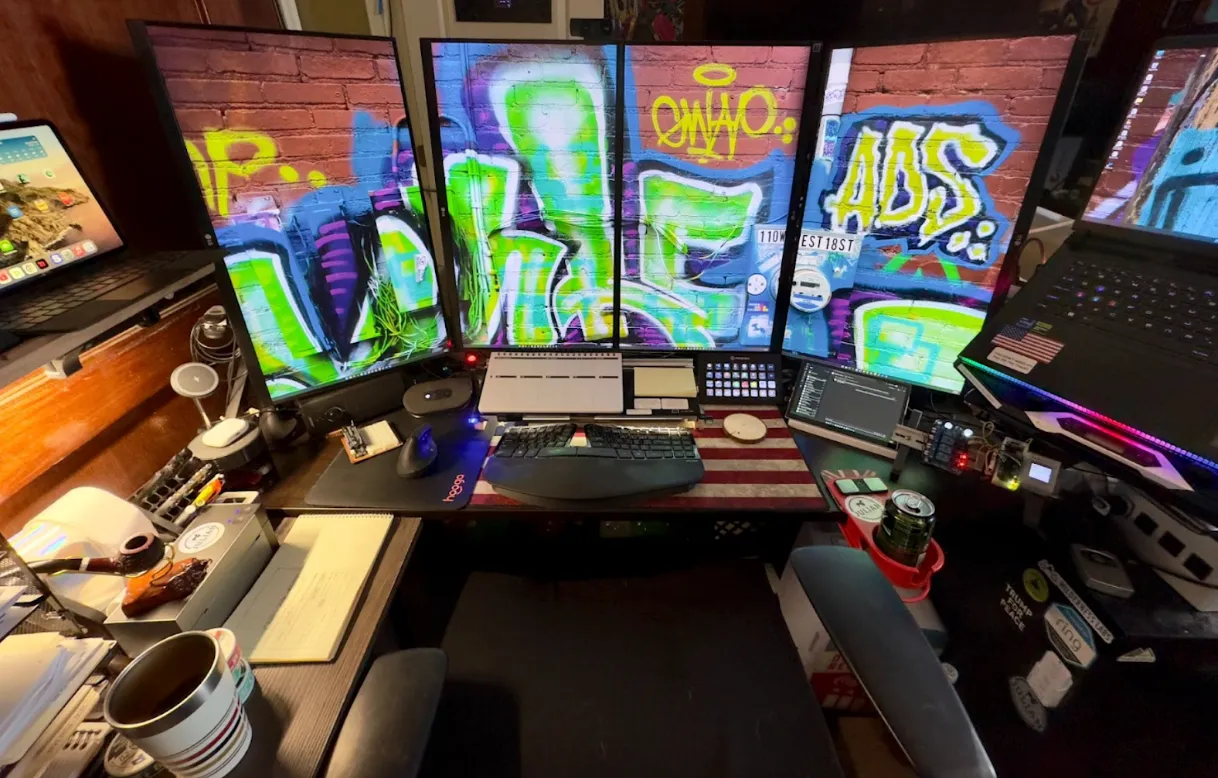 A computer workstation with four vertical monitors displaying graffiti art, surrounded by various devices, keyboards, and office supplies.