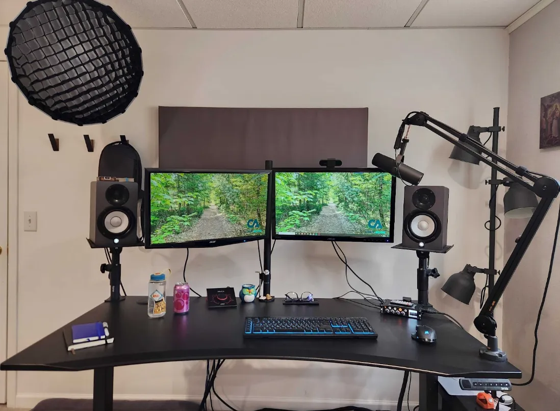 Dual-monitor computer desk setup with keyboard, mouse, speakers, microphone on adjustable arm, water bottles, and various desk accessories in a minimalist room.