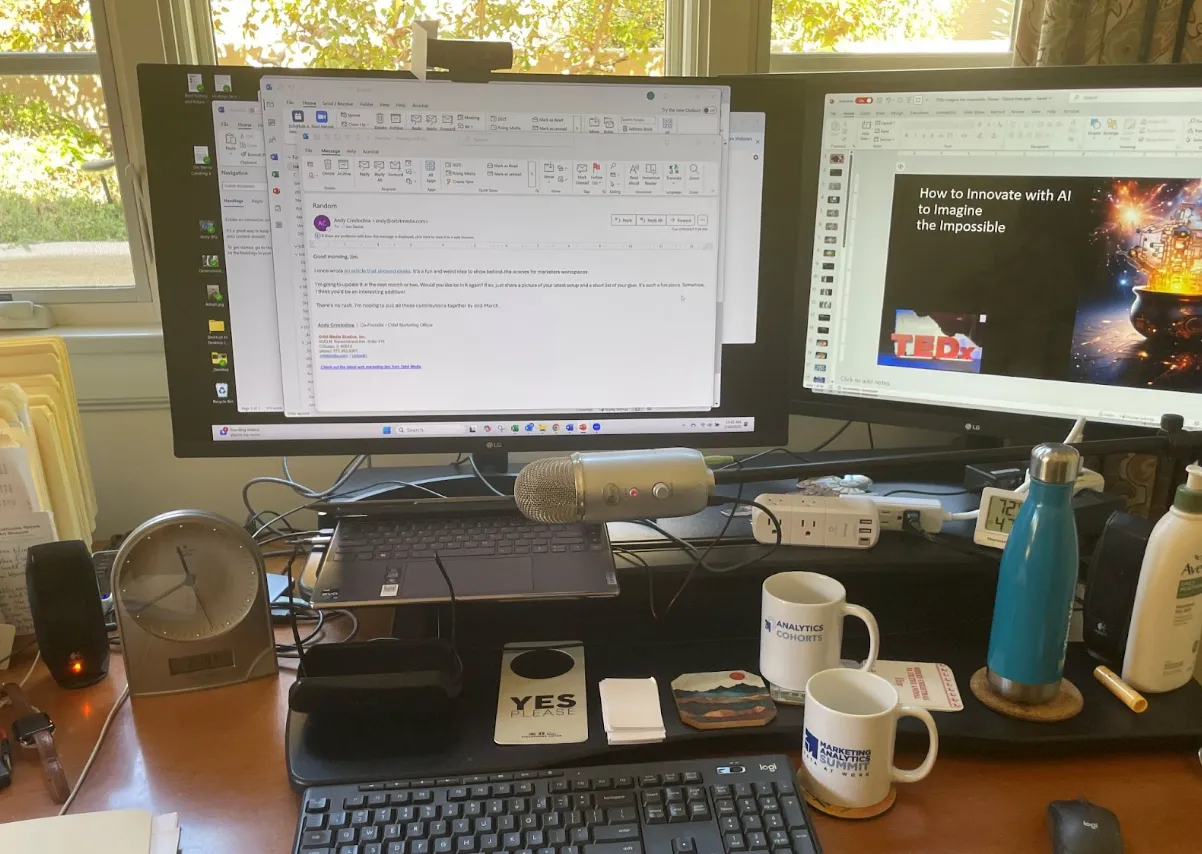 Home office desk setup with dual monitors displaying an email and a TEDx presentation, microphone, keyboard, mugs, water bottles, clock, and various office supplies.