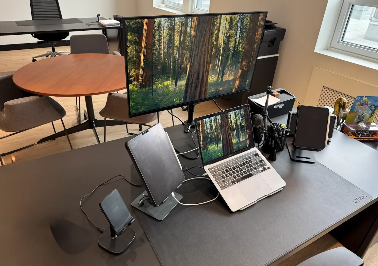 A modern office desk setup with a laptop, tablet, smartphone stand, monitor displaying a forest, and various office supplies; round table and chairs in the background.
