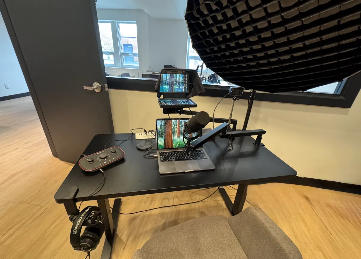 A podcast recording setup featuring a laptop, microphone on an adjustable arm, audio interface, headphones, tablet, and camera on a black desk in a modern room.