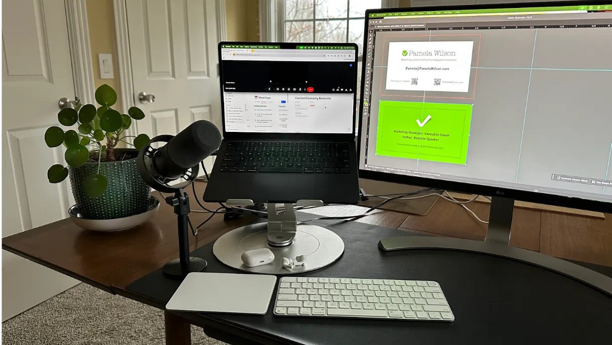 A home office setup with a laptop on a stand, external monitor, keyboard, trackpad, microphone, AirPods, and a potted plant on a wooden desk.