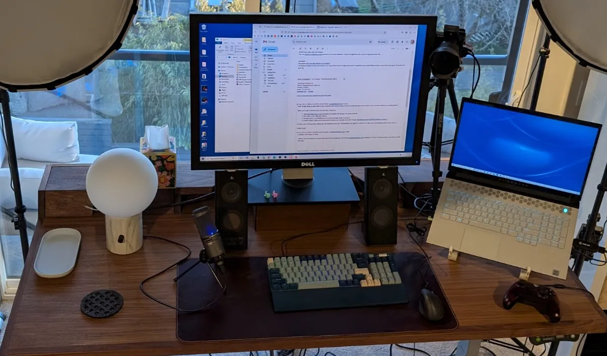 A desk setup with a large monitor displaying text, a mechanical keyboard, microphone, speakers, lamp, closed laptop, game controller, and various accessories. A window is in the background.