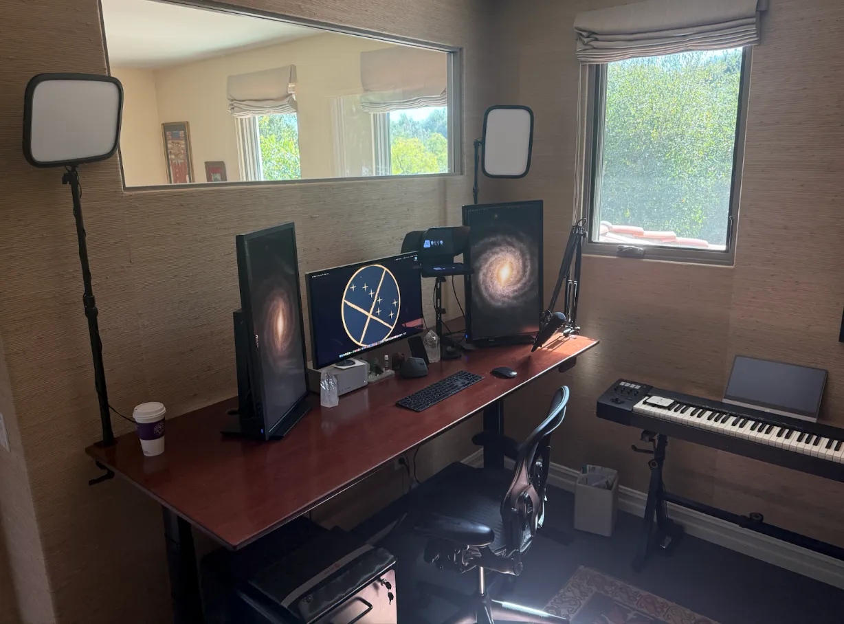 A home office setup with a dual-monitor computer, desk chair, microphone, two lights, coffee cup, window view, and an electronic keyboard against the wall.