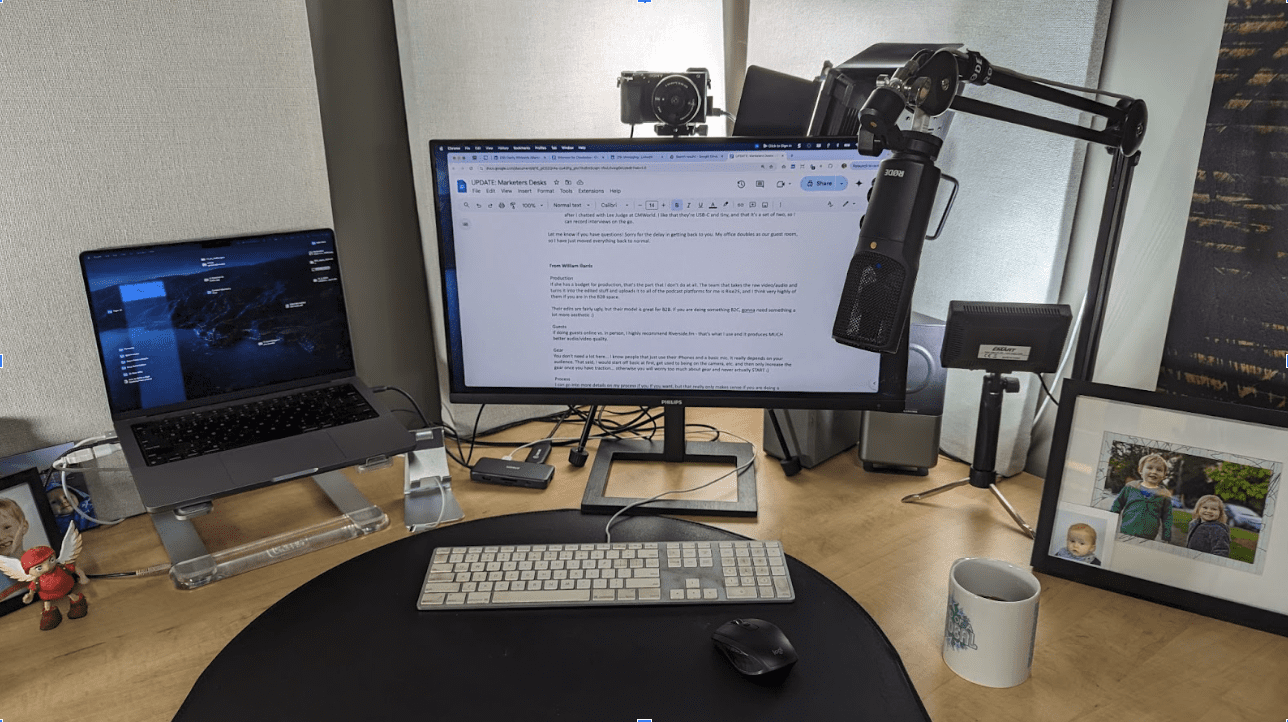 A home office setup with a laptop, external monitor displaying text, microphone on a boom arm, keyboard, mouse, camera, framed photo, mug, and small figurines on a wooden desk.