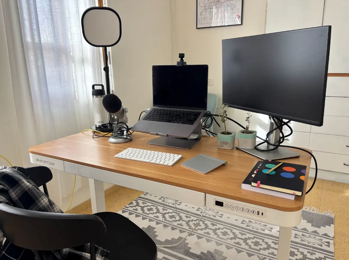 A modern desk setup with a laptop on a stand, external monitor, keyboard, mouse, microphone, ring light, notebooks, potted plants, and a chair in a well-lit room.