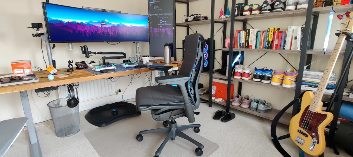 A home office setup with a large monitor, office chair, desk accessories, bookshelf with books and shoes, a guitar, and various personal items.