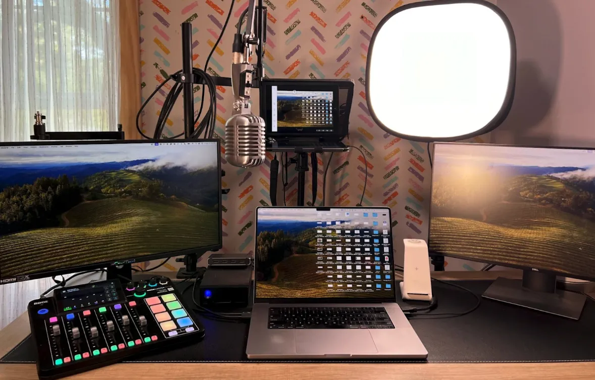 A home office setup with three monitors, a laptop, a podcast mixer, a microphone, and a large ring light on a desk.