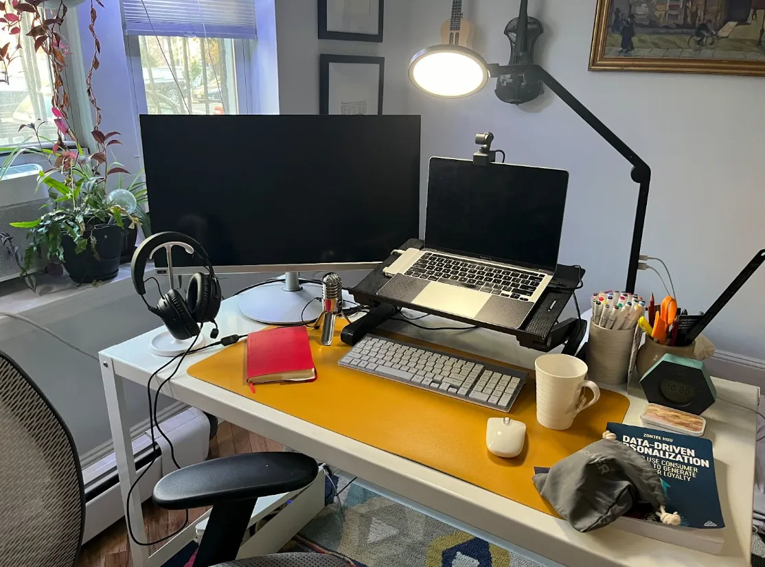 A home office desk with a monitor, laptop on a stand, keyboard, mouse, headset, lamp, notebook, pens, coffee mug, and a data analytics book. Plants and wall art in the background.