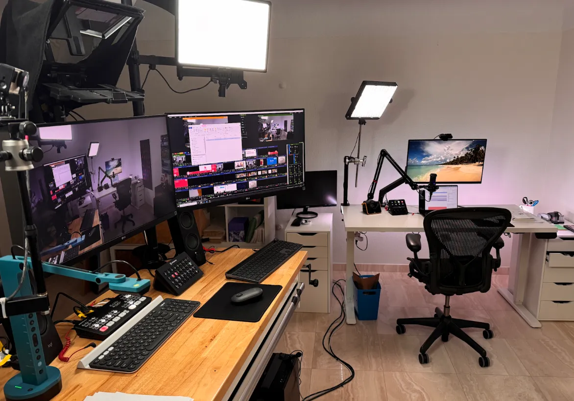 A home office setup with multiple monitors, video production equipment, studio lights, microphones, and ergonomic chairs arranged at two desks.