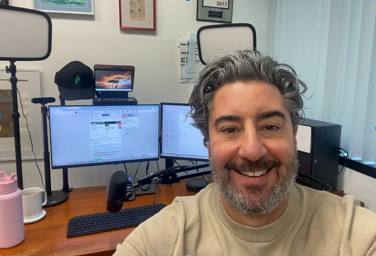 A person smiling at their desk with dual monitors, a microphone, water bottle, and lighting equipment in a home office setting.