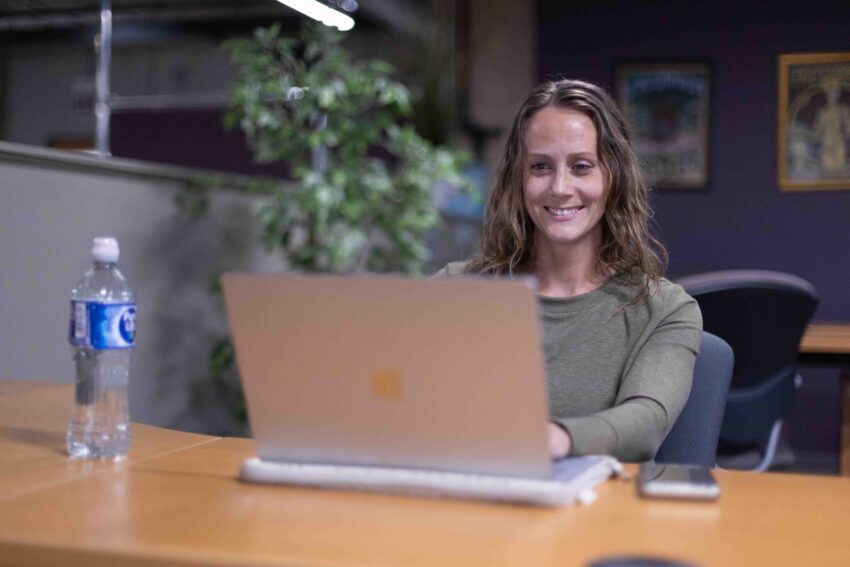 A woman sits at a desk using a laptop, with a water bottle and smartphone nearby, in an office setting.