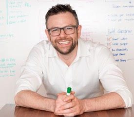 Man with glasses and beard, wearing a white shirt, sits at a table holding a green marker, smiling, with a whiteboard and notes in the background.
