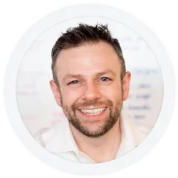 A man with short brown hair and a beard smiles at the camera, wearing a white shirt, with a blurred white background.