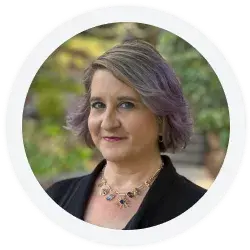 Woman with short, wavy, light brown and purple hair wearing a dark blazer and a beaded necklace, standing outdoors with greenery in the background.