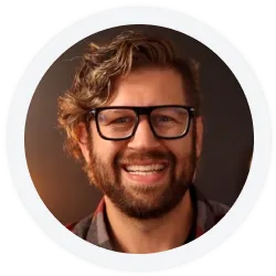 A man with wavy hair, a beard, and glasses smiles at the camera against a dark background.