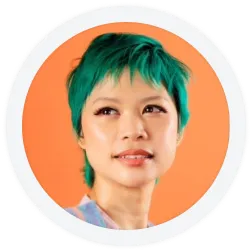 Person with short, bright teal hair and natural makeup, wearing a light-colored top, looks slightly upward against an orange background.